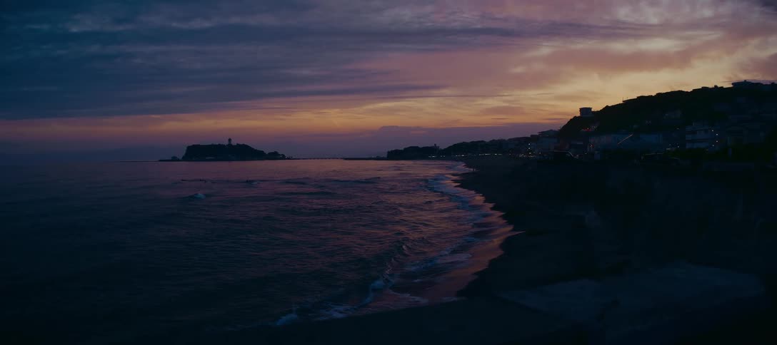 A tranquil view of the ocean waves washing ashore along the coastline of a town at dusk under a colorful purple and orange sky