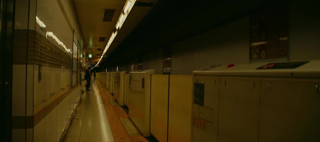 A lone person stands still at the far end of a long, empty, and dimly lit subway station
