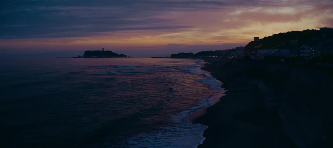 A serene wide shot of the Japanese coastline at dusk shows gentle waves washing ashore against a beautiful purple and orange sunset sky