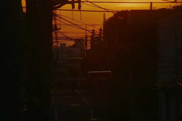 A silhouetted cyclist rides through a quiet Japanese street intersection filled with power lines during a vibrant orange sunset