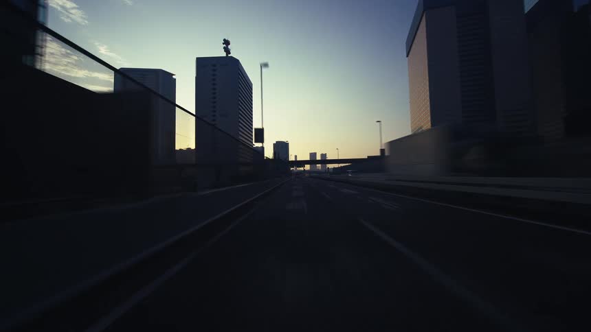 A stylized point-of-view shot speeds along a deserted city highway, surrounded by towering skyscrapers silhouetted by the rising sun