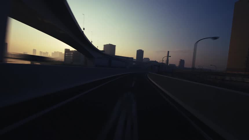 A dynamic point-of-view timelapse captures a drive along a modern city highway under massive overpasses during a beautiful sunset