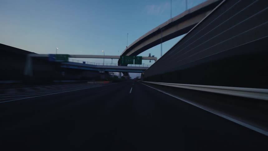 A point-of-view shot of a car driving on a nearly empty urban expressway under and around concrete overpasses at dawn