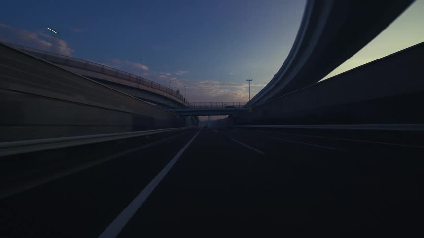 A point-of-view timelapse captures a fast drive along an urban expressway under complex overpasses at dawn