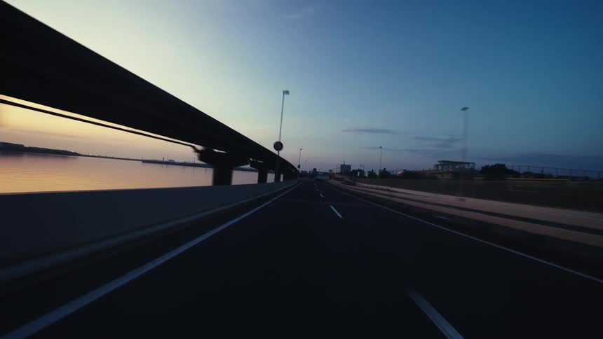 A smooth point-of-view shot driving on a highway overpass next to a body of water during a beautiful twilight sunset