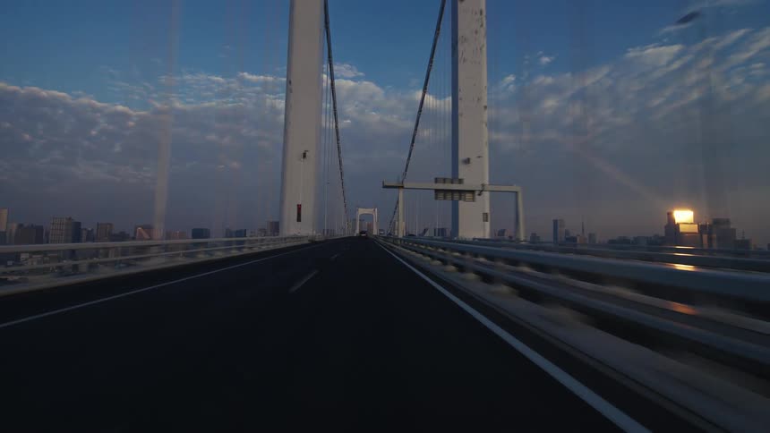 A point-of-view shot driving across the iconic Rainbow Bridge in Tokyo at sunrise, with the city skyline in the background