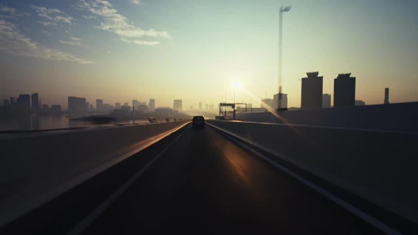 A stylized, rear-facing timelapse shows a fast drive over a modern city bridge towards the rising sun
