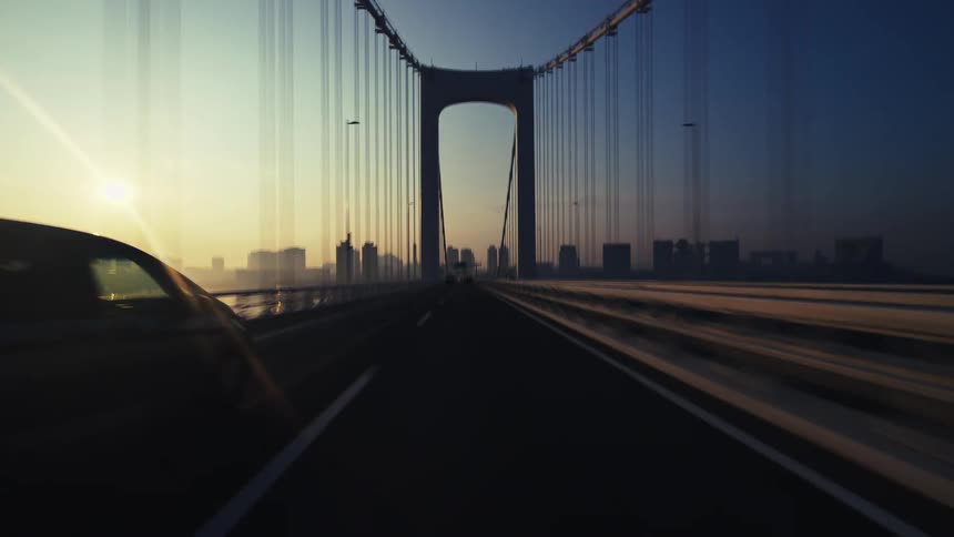 A cinematic point-of-view shot driving across a large suspension bridge toward a city skyline at sunrise