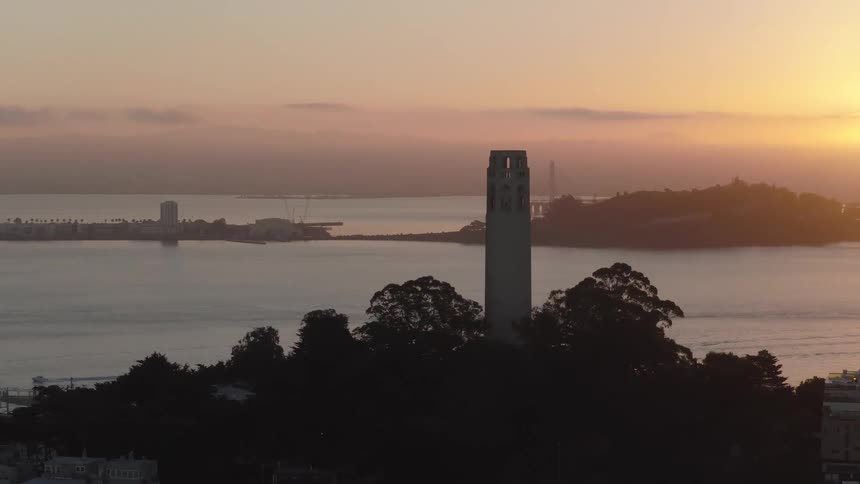 Aerial view of San Francisco's Coit Tower silhouetted against a hazy sunrise, with Treasure Island and the Bay Bridge in the distance