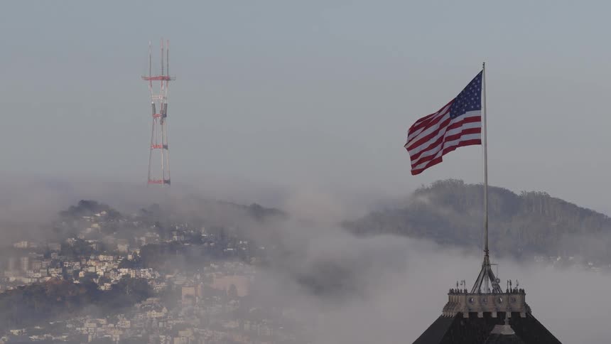 An American flag waves slowly in the wind with the iconic Sutro Tower and foggy San Francisco hills in the background