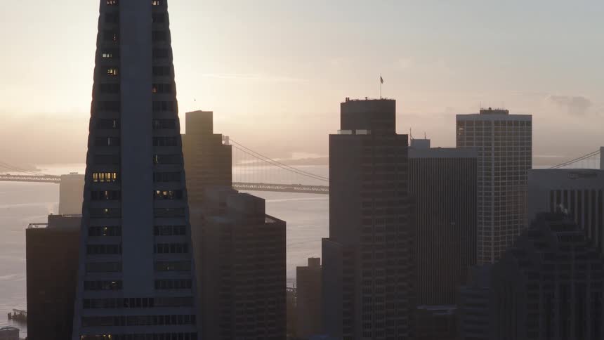Aerial view of the San Francisco skyline, featuring the Transamerica Pyramid and the Bay Bridge in the background during a hazy sunrise