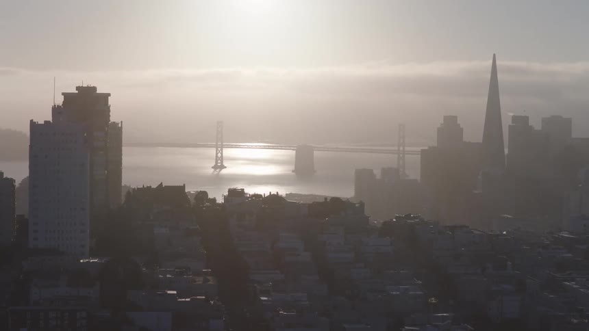 A hazy sunrise illuminates the San Francisco skyline, highlighting the Bay Bridge and Transamerica Pyramid