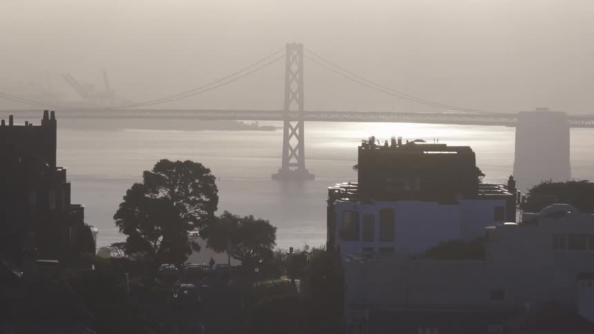 A hazy sunrise view of the San Francisco Bay Bridge with silhouetted city buildings in the foreground