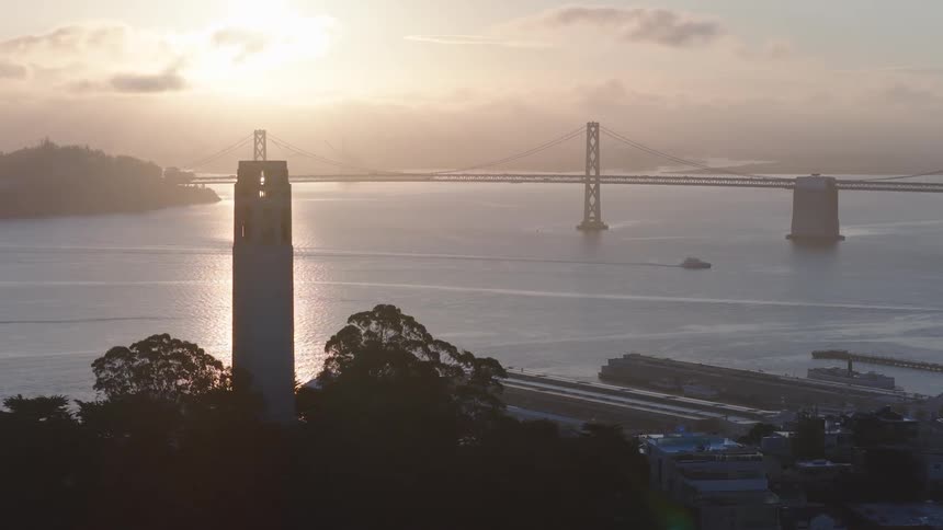 A serene wide shot of San Francisco's Coit Tower and Bay Bridge silhouetted against a beautiful golden sunrise over the bay