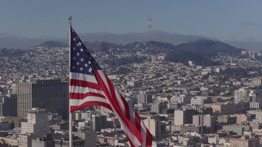 The American flag waves in slow motion over the San Francisco, California skyline, with the Sutro Tower visible in the distance