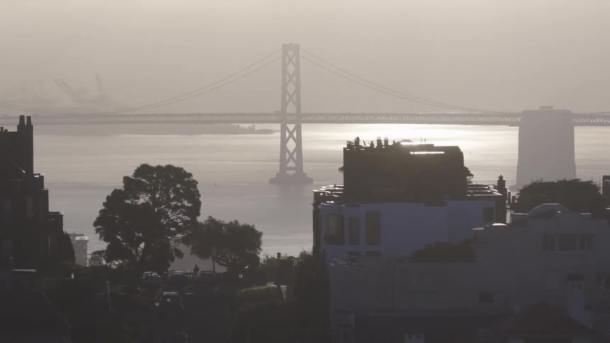 A hazy sunrise over the San Francisco Bay reveals the Bay Bridge with silhouetted city buildings in the foreground
