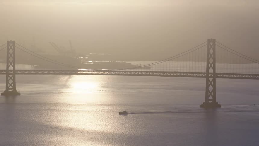 The San Francisco Bay Bridge is silhouetted against a golden, hazy sunrise as a boat crosses the shimmering water