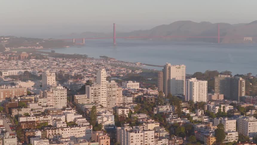 An aerial view slowly drifts over the city of San Francisco towards the Golden Gate Bridge during a hazy golden hour sunrise