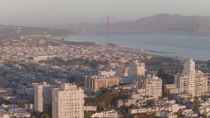 A slow aerial view moves across the sunlit buildings of a San Francisco neighborhood, with the iconic Golden Gate Bridge and bay in the hazy background