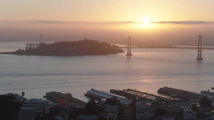 A serene aerial shot captures the sunrise over San Francisco Bay, silhouetting Coit Tower, Treasure Island, and the Bay Bridge against a glowing sky