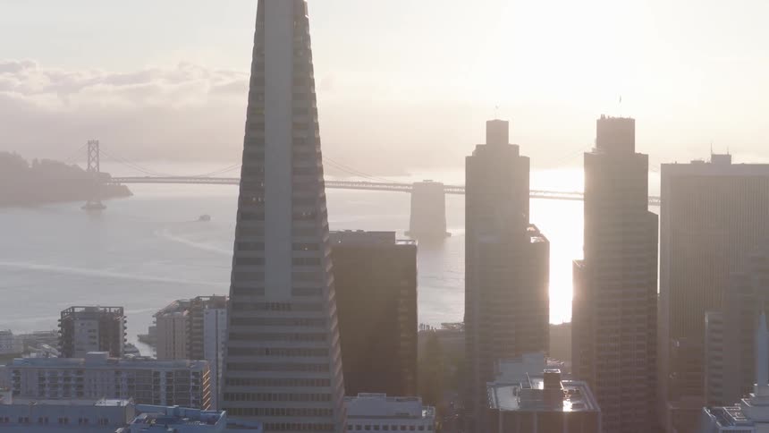 A slow aerial shot pulls back from the San Francisco skyline, revealing the Transamerica Pyramid and the Bay Bridge during a hazy sunrise