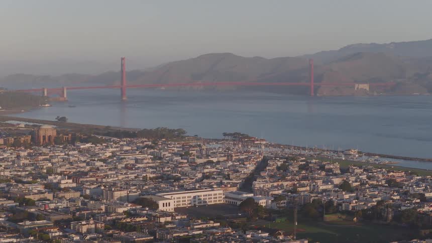 A wide, static shot captures the iconic Golden Gate Bridge behind the San Francisco cityscape and marina during a calm, hazy morning