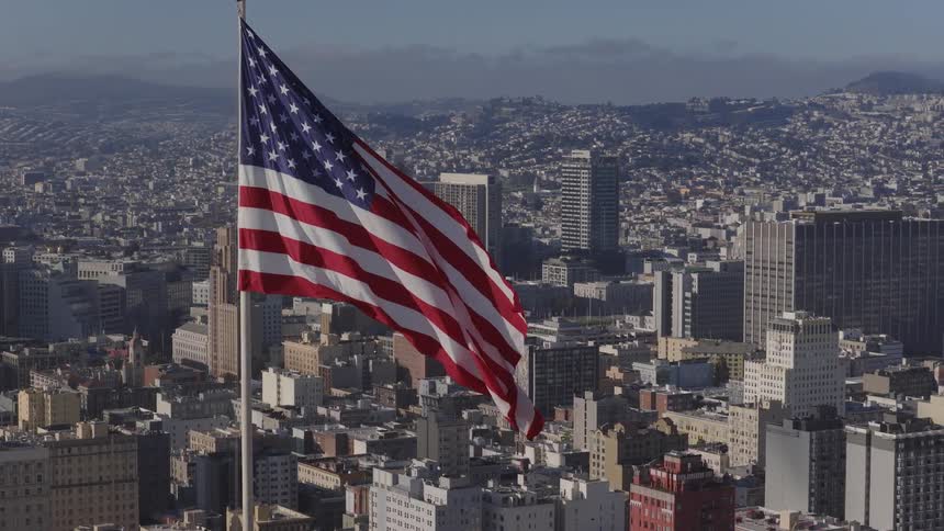 A majestic aerial shot captures the American flag waving in slow motion over the sunny San Francisco cityscape