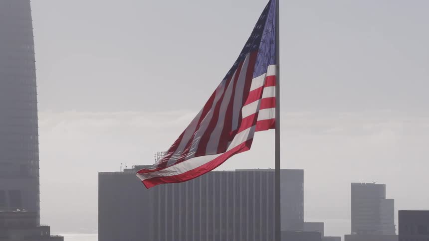 A large American flag waves majestically in slow motion against the hazy San Francisco city skyline
