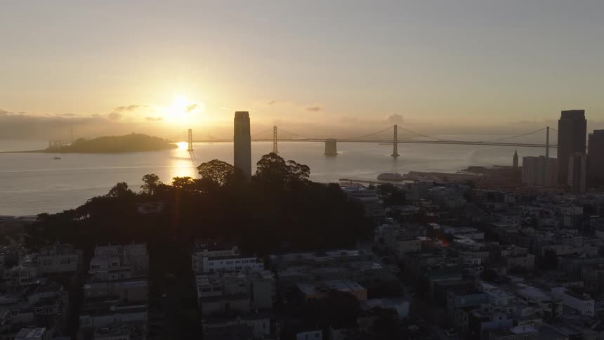 A serene aerial view of San Francisco at sunrise, featuring the silhouetted Coit Tower and the majestic Bay Bridge glowing in the morning light