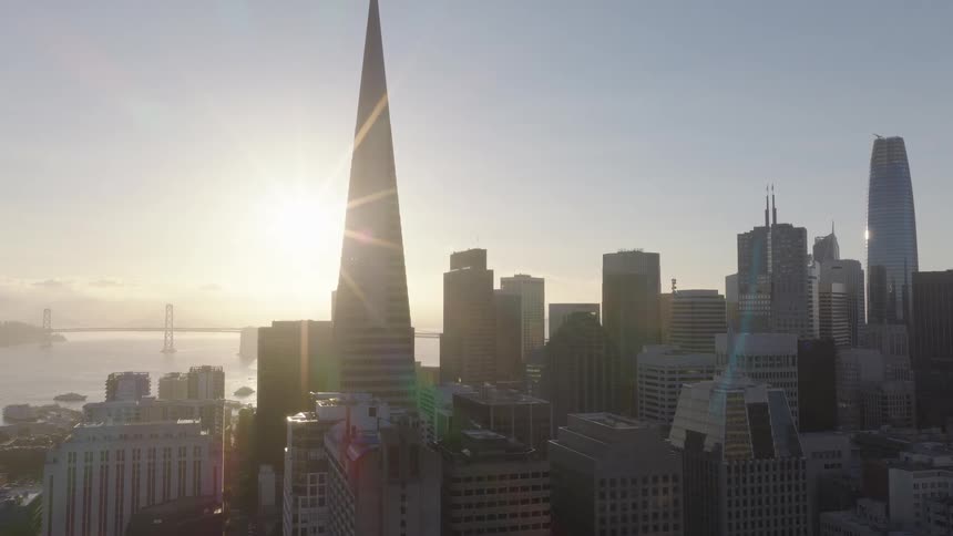 A slow aerial pullback reveals the iconic San Francisco skyline at sunrise, with a bright sunburst behind the Transamerica Pyramid