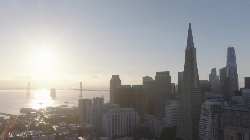A hazy morning sun rises over the iconic San Francisco skyline, featuring the Transamerica Pyramid and the Bay Bridge in the distance