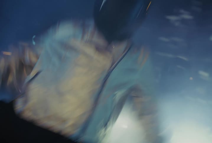 A person in a silver jacket and a black helmet dances energetically in a crowd at a smoky, futuristic night party