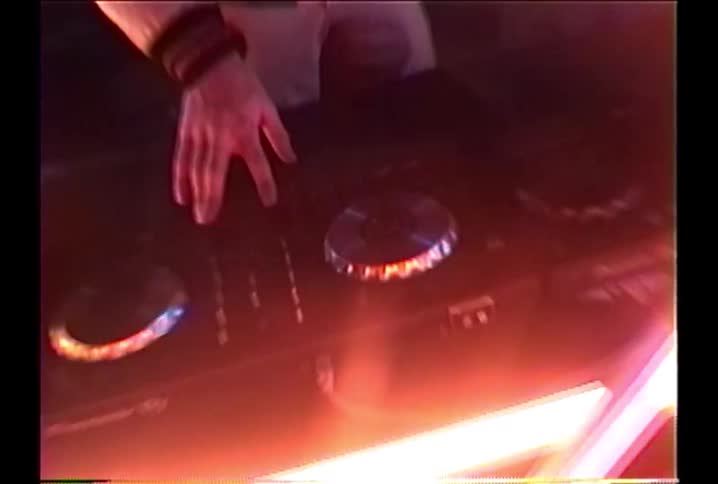A retro VHS-style overhead shot zooms in on a DJ's hands mixing music on a turntable in a club with red neon lights