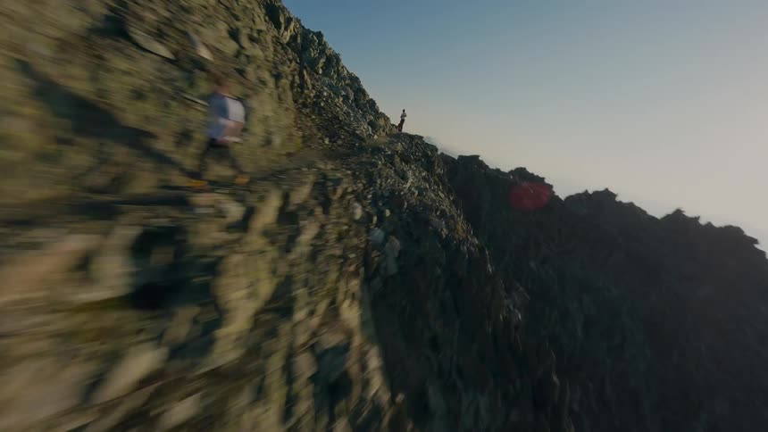 A thrilling FPV drone shot soars dangerously close to a rugged mountain ridge, tracking adventurers as they hike a narrow path at high altitude