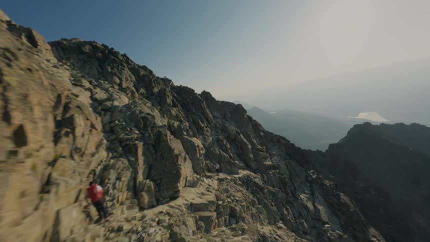 A dynamic aerial shot follows two hikers as they ascend a narrow, rocky path along a steep mountain ridge