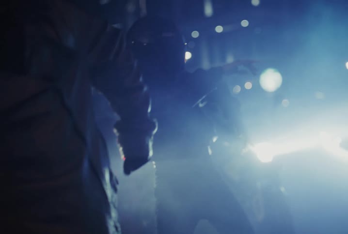 A silhouetted figure in a motorcycle helmet dances at a vibrant, smoke-filled party with bright blue lights