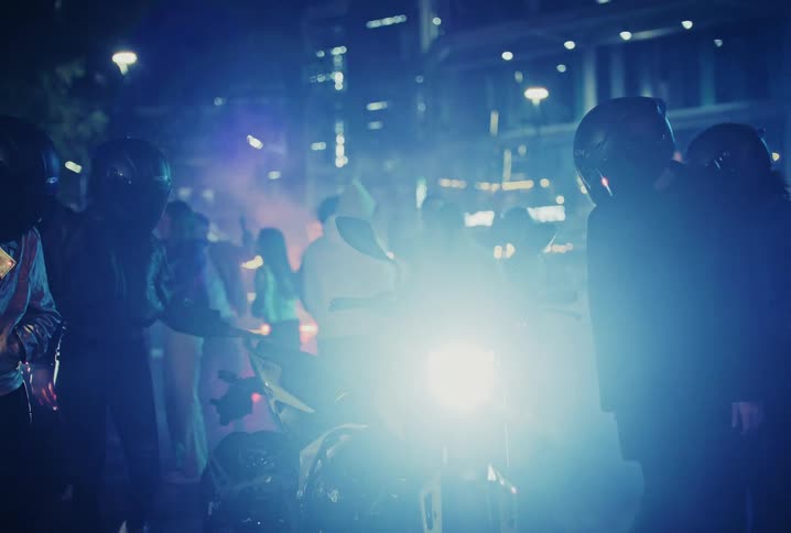 Bikers hangout at night in the city, illuminated by a motorcycle's bright headlight and surrounded by blue smoke