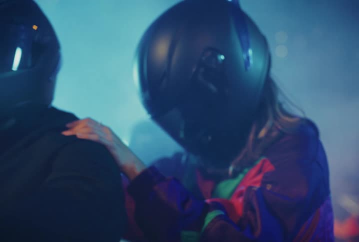 A woman in a retro jacket puts on her helmet and holds on to a man as they get ready for a motorcycle ride in a smoky, blue-lit environment