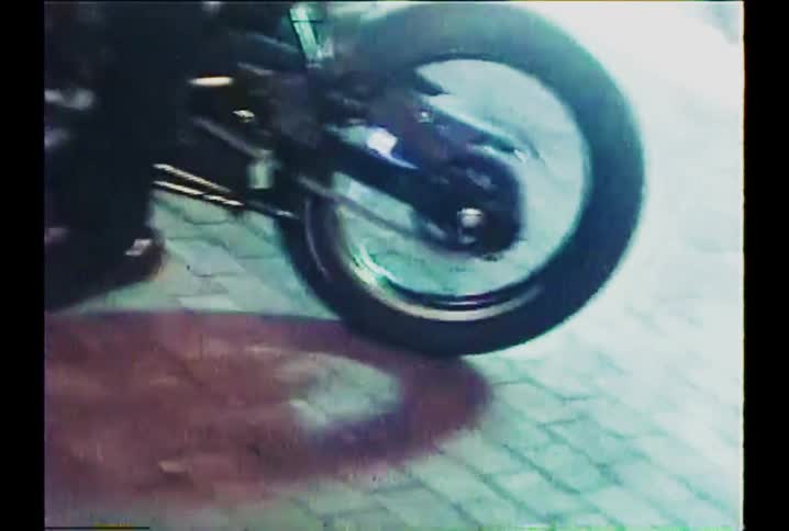 Distorted VHS-style footage zooms in on a motorcycle's back wheel spinning on a brick street at night