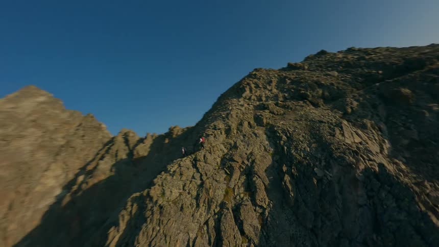 A breathtaking FPV drone shot follows two adventurers hiking up a steep, rocky mountain summit during a beautiful sunrise