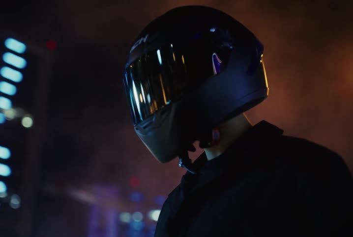 A mysterious person in a motorcycle helmet emerges from the smoky, neon-lit city night in a slow-motion reveal