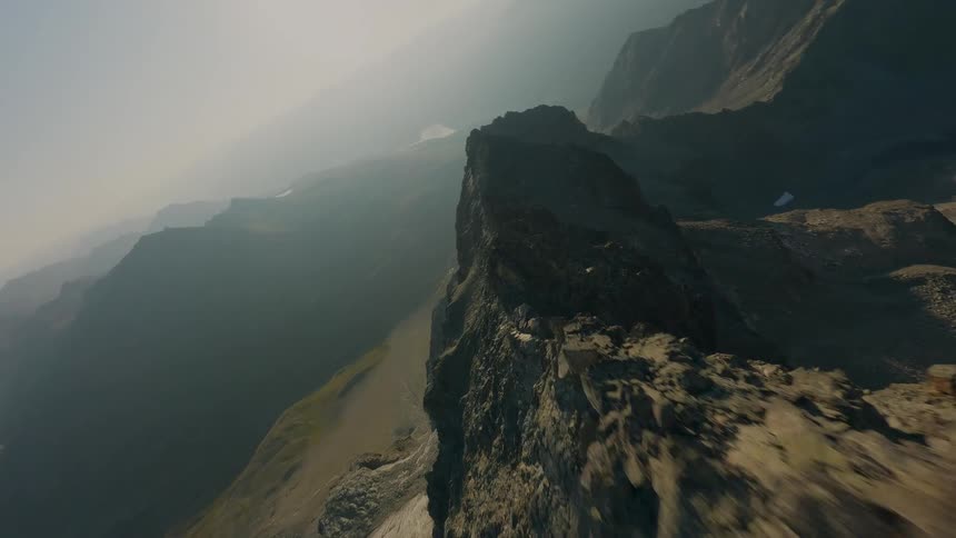 A dramatic FPV drone shot skims the sharp edge of a mountain ridge, revealing a vast, hazy mountainscape