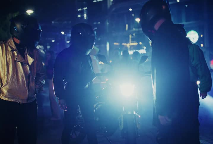 A group of mysterious bikers wearing helmets gather around a motorcycle with its bright headlight on a neon-lit city street