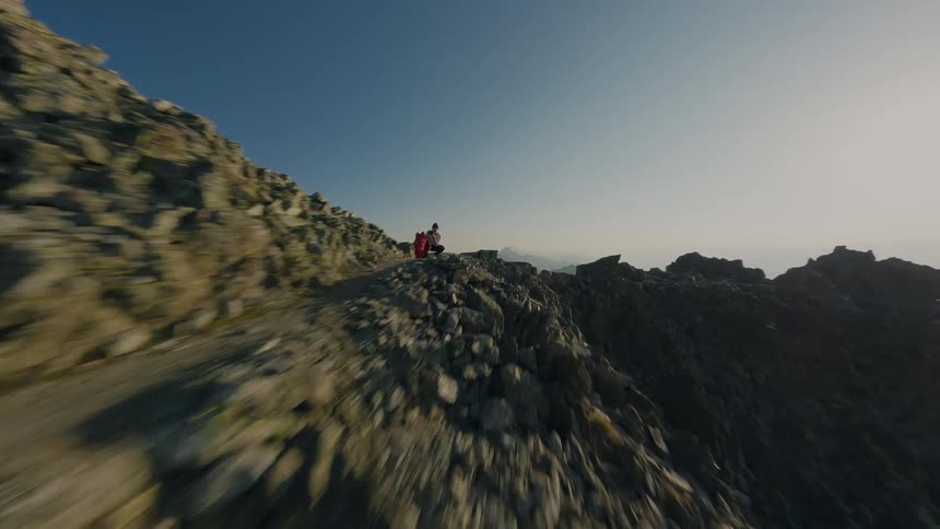 An FPV drone speeds along a rocky mountain ridge, flying past a lone hiker to reveal a stunning panoramic view of a vast mountain range