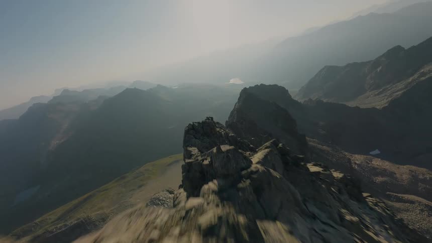An FPV drone soars along a dramatic and rocky mountain ridge, following a lone hiker towards the summit on a hazy, sunlit day