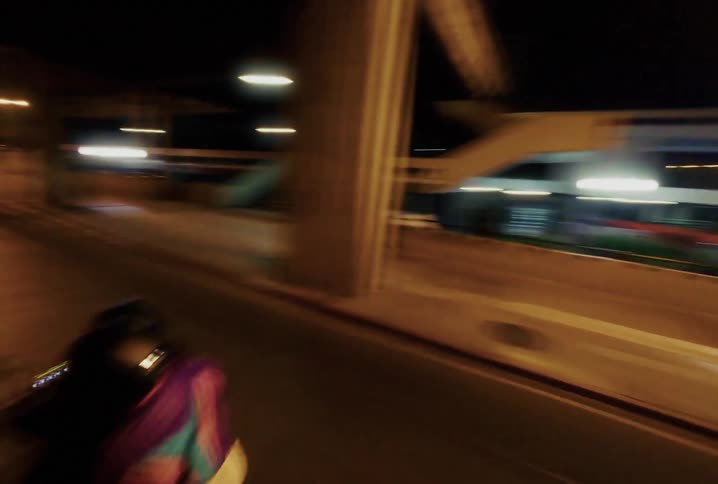 A dynamic, moving camera follows a couple riding a motorcycle through the city streets at night