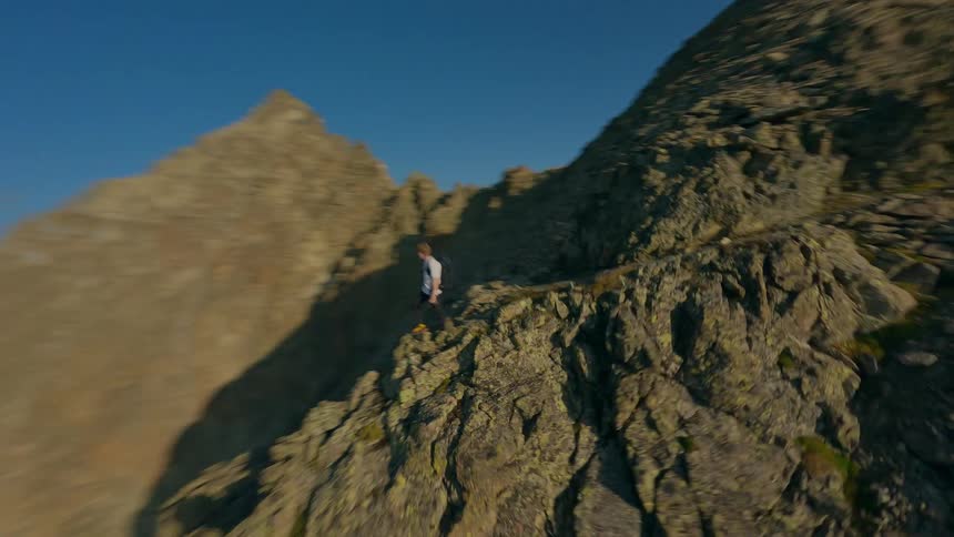 Dynamic FPV drone shot orbits a lone hiker standing on a spectacular, rocky mountain ridge during a hazy sunrise