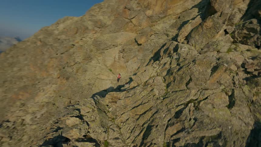 Exhilarating FPV drone footage follows two mountaineers as they carefully climb a narrow, rocky ridge high in the sunlit mountains
