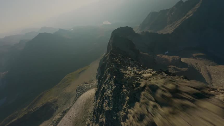 A fast FPV drone flight skims along a sharp, rocky mountain ridge during a hazy sunrise, revealing an epic, sprawling alpine vista