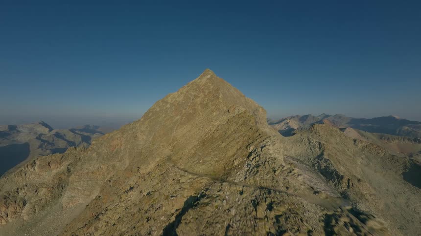 A breathtaking FPV drone flight along a treacherous rocky ridge towards a majestic mountain summit under a clear blue sky